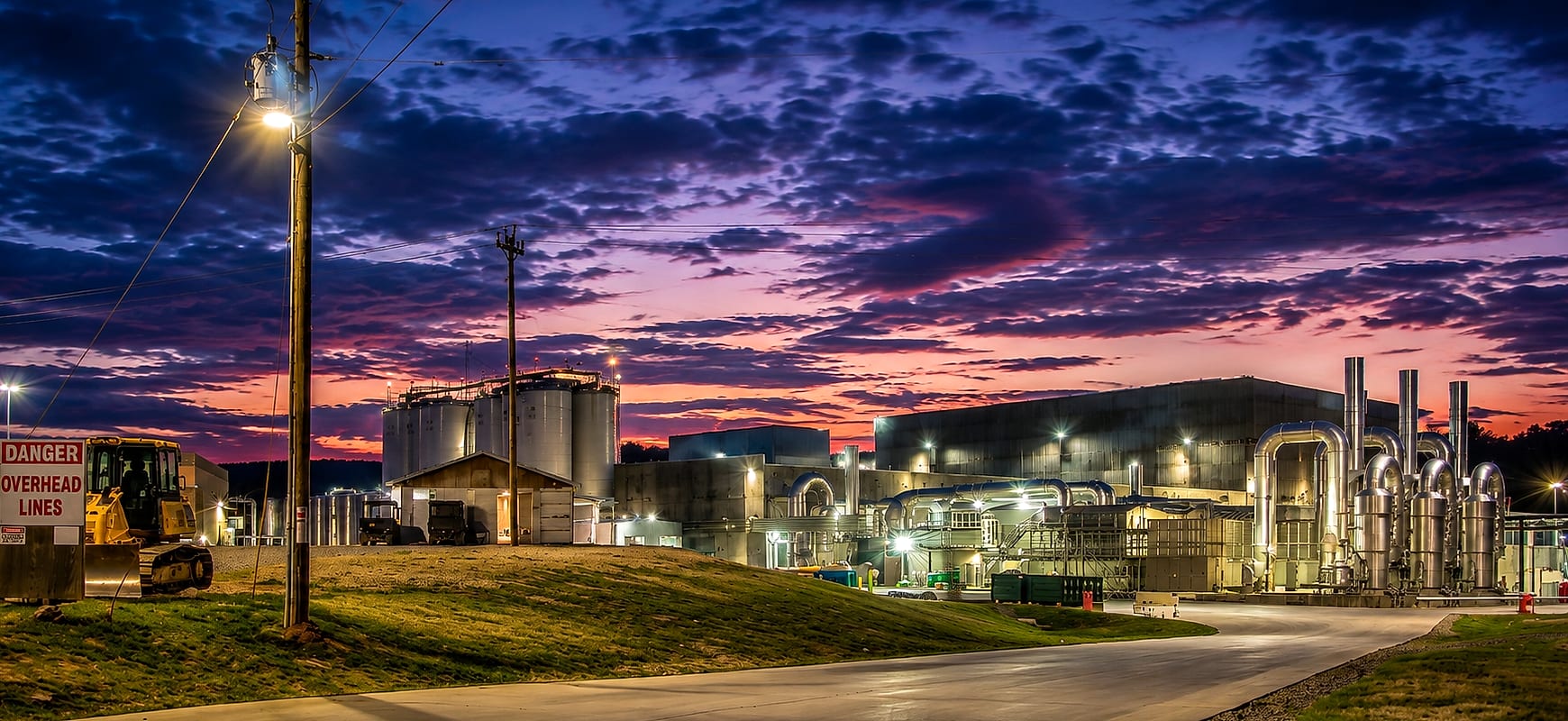 Industrial process facility at dusk with storage silos, exhaust stacks and process equipment lit against an evening sky