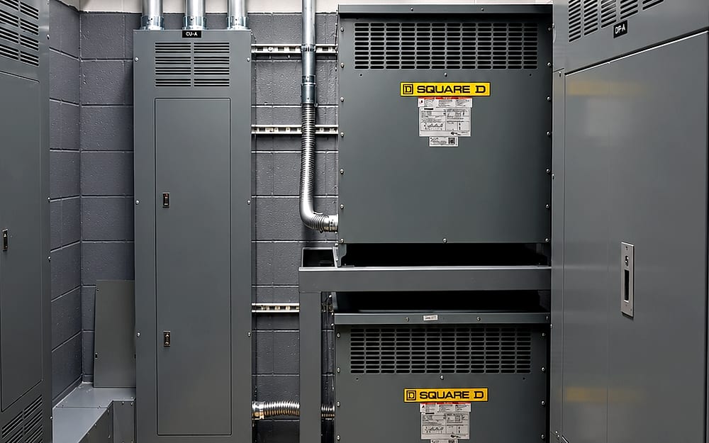 Electrical room with dry-type transformers, distribution and lighting panels identified by section, flexible conduit whips and yellow working-clearance floor marking